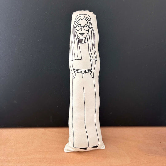 Screen printed doll cushion with white fabric and black illustration depicting feminist activist Gloria Steinem with long hair, round glasses, a polo neck jumper and her hands in her pockets