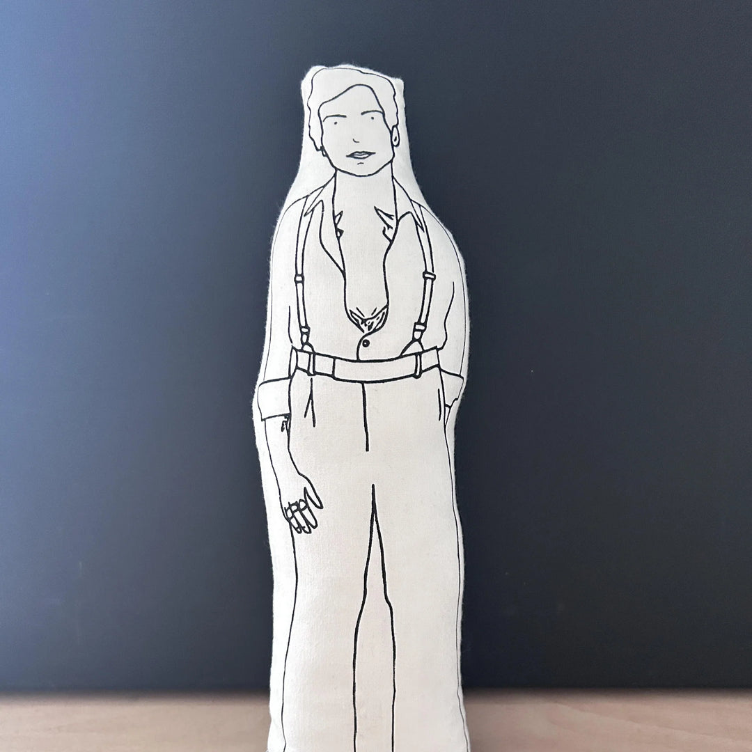 Screen printed doll cushion with white fabric and black illustration depicting singer-songwriter Harry Styles wearing trousers with braces and an undone shirt showing chest tattoos