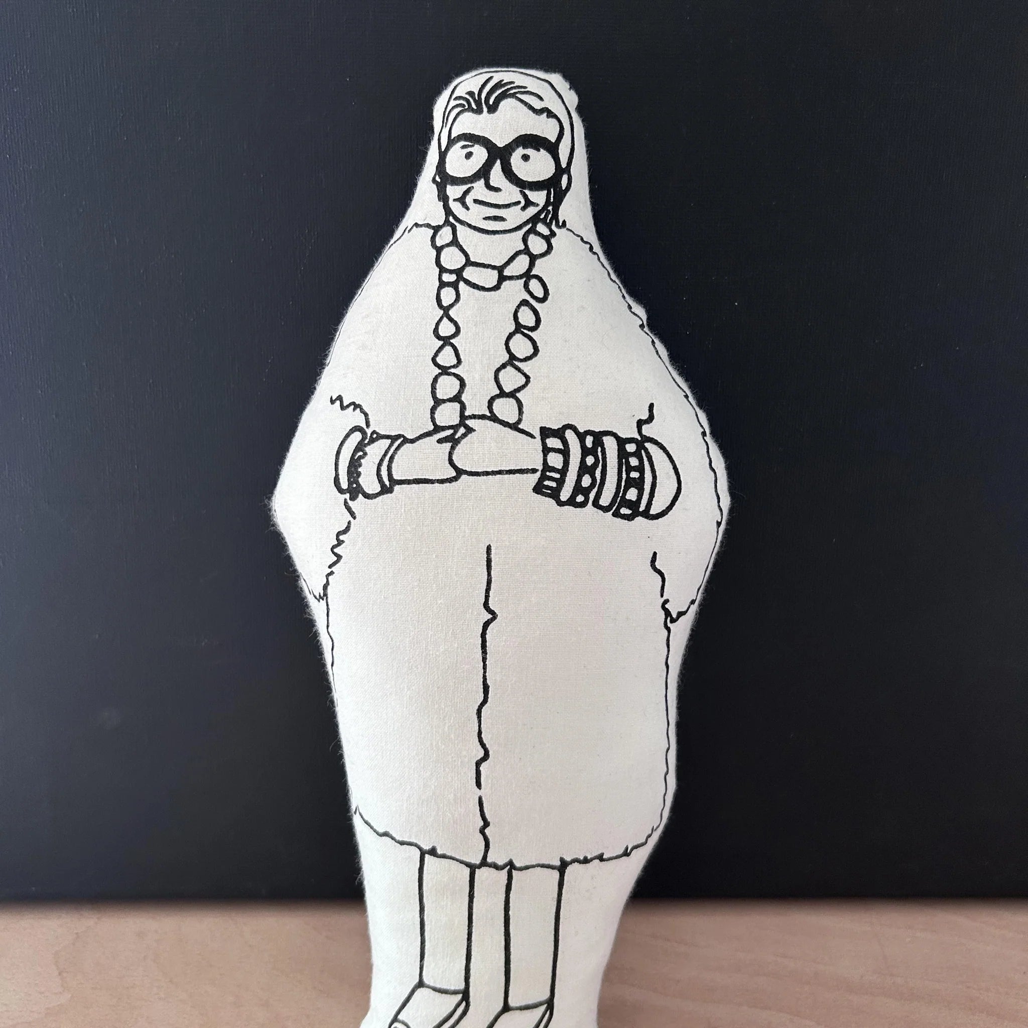 Screen printed doll cushion with white fabric and black illustration depicting fashion designer Iris Apfel wearing a big fluffy coat, large statement jewellery and round glasses