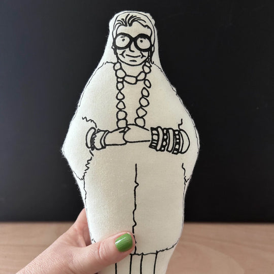 Hand holding screen printed doll cushion of Iris Apfel against a dark background