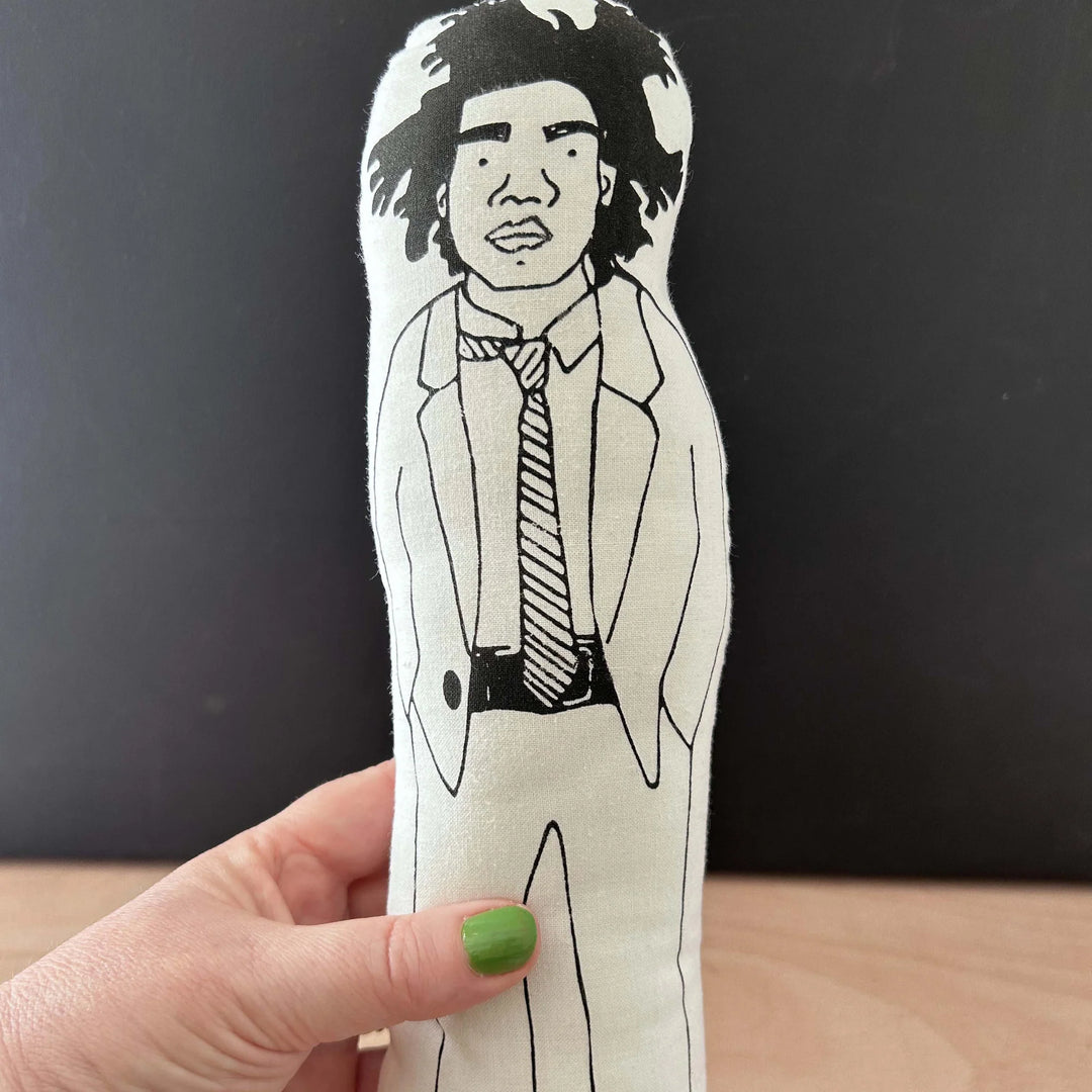 A hand holding screen printed doll cushion of artist Jean Michel Basquiat against a dark background