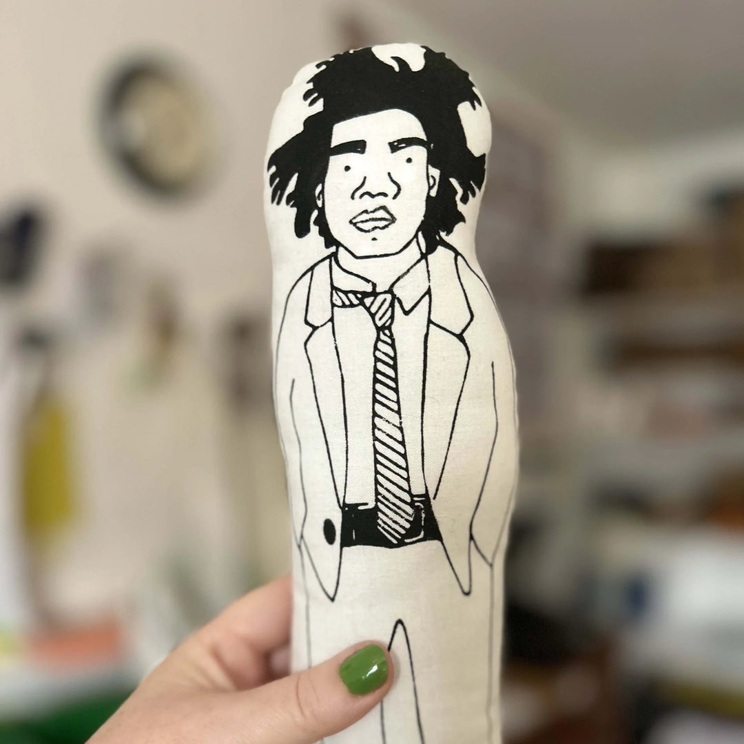 Hand holding screen printed doll cushion of artist Jean Michel Basquia against a blurred background
