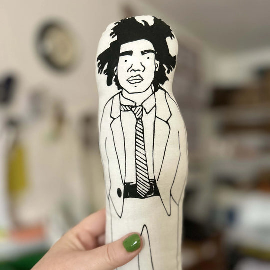 Hand holding screen printed doll cushion of artist Jean Michel Basquia against a blurred background