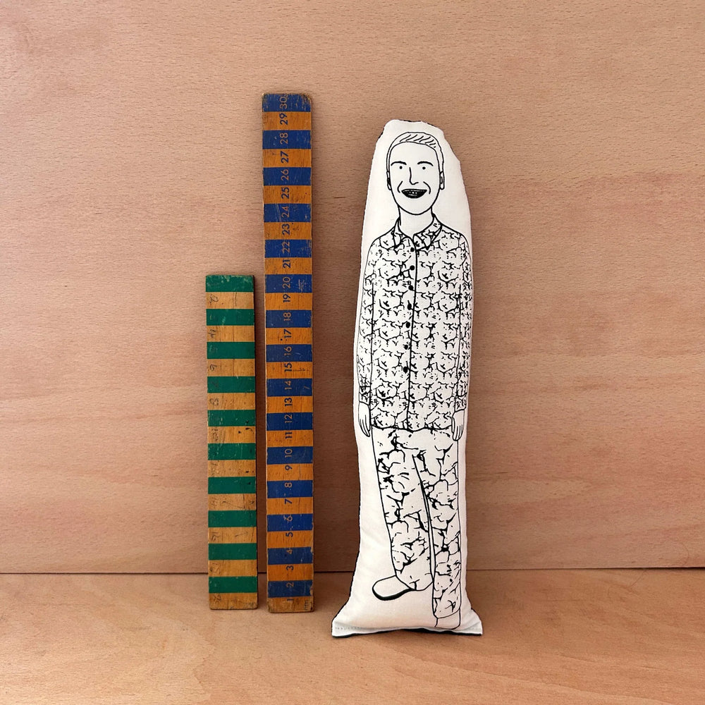 Screen printed doll cushion of Joe Lycett next to two wooden rulers on a wooden background