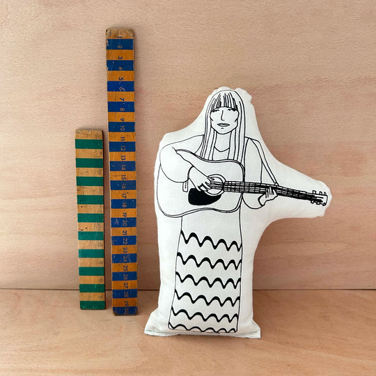 Screen printed doll cushion of singer-songwriter Joni Mitchell playing a guitar next to two wooden rulers