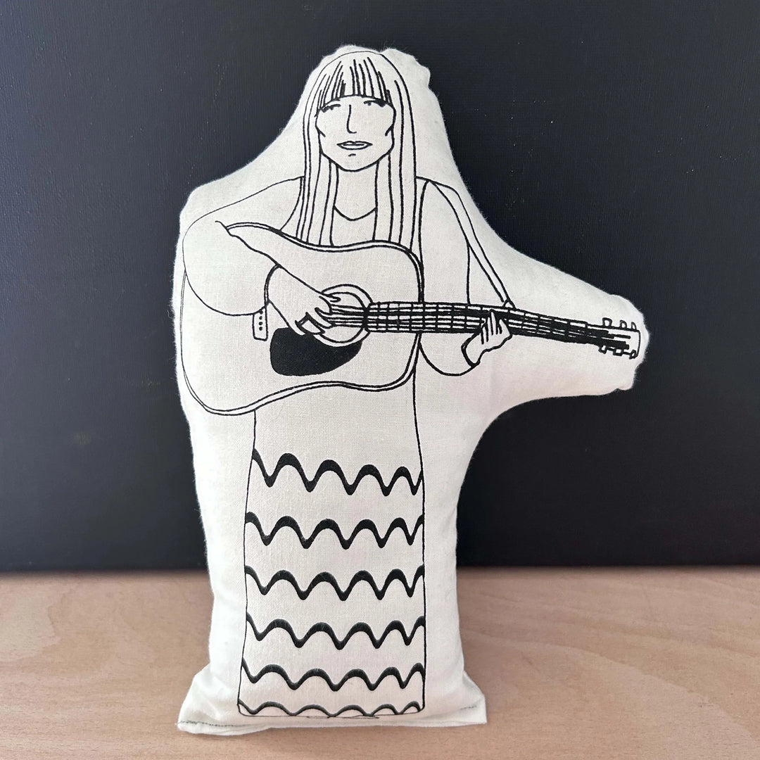Screen printed doll cushion with white fabric and black illustration depicting siner Joni Mitchell playing a guitar wearing a long patterned dress