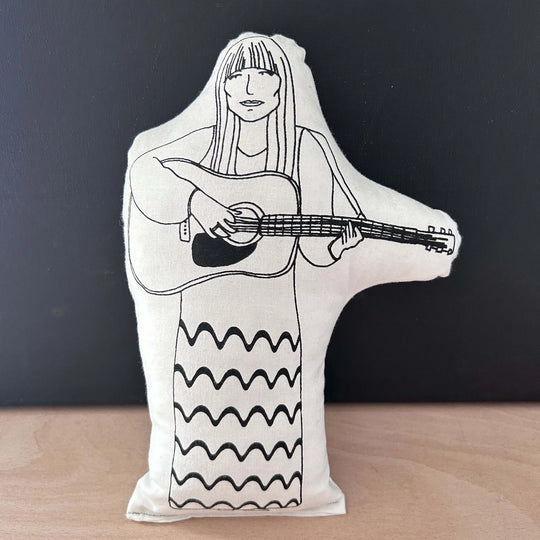 Screen printed doll cushion with white fabric and black illustration depicting siner Joni Mitchell playing a guitar wearing a long patterned dress