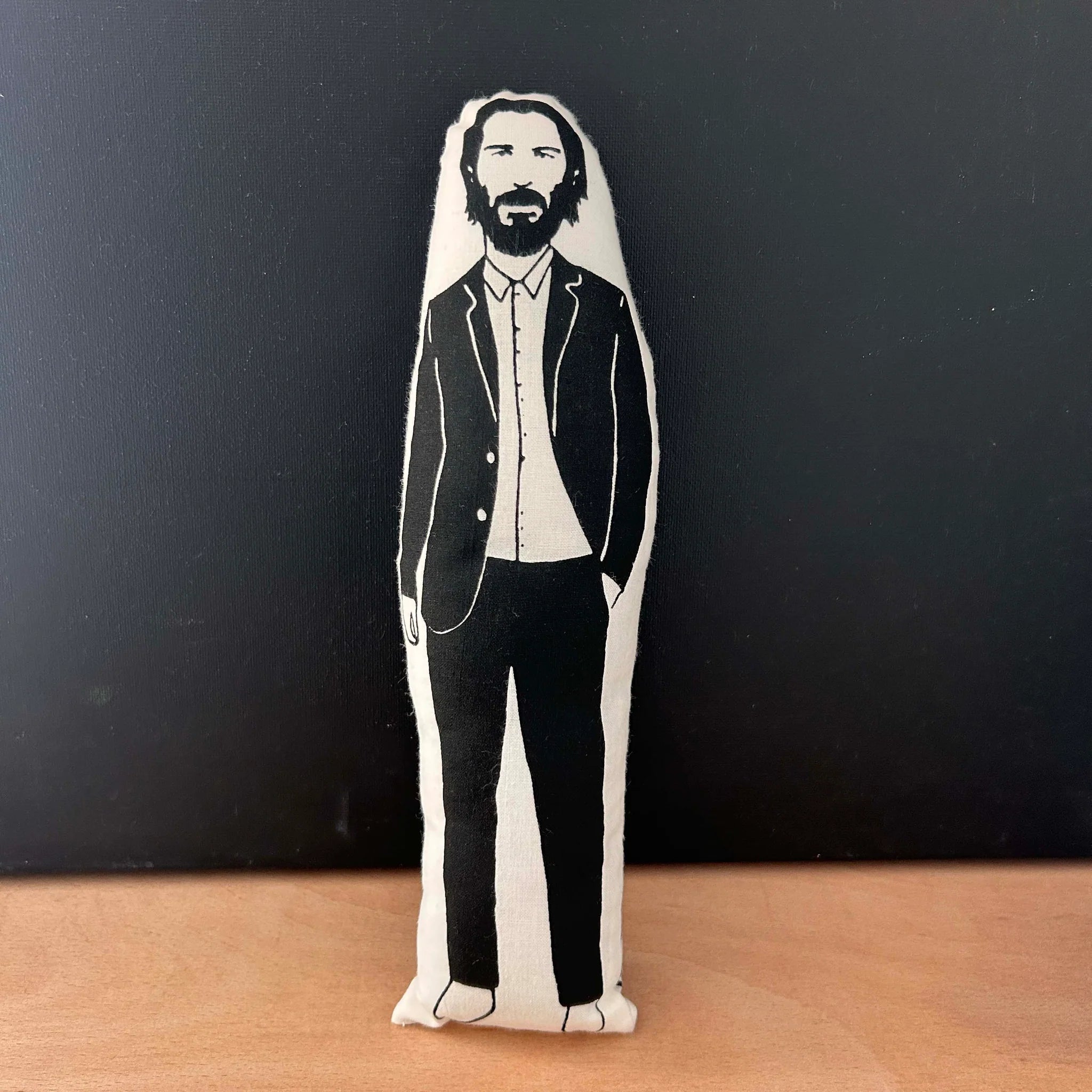 Screen printed doll cushion with white fabric and black illustration depicting actor Keanu Reeves wearing a casual black suit with one hand in his pocket