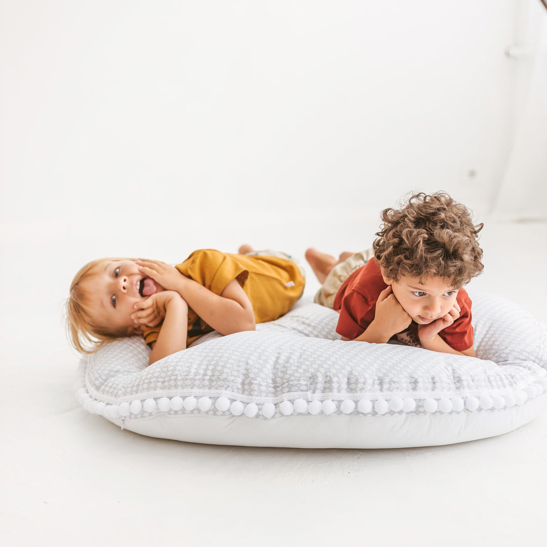 Two children lying on a round light grey floor cushion with pom-pom trim against a white background