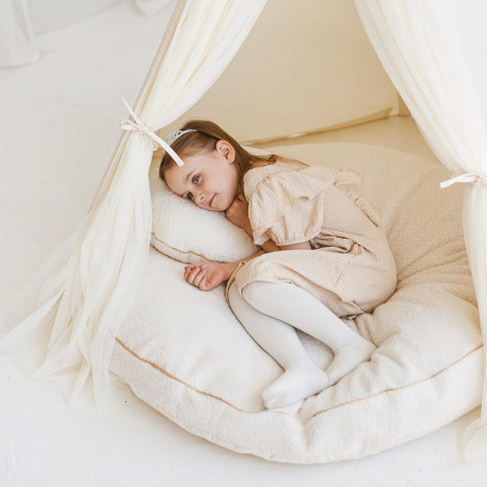 Child lying on pale beige floor cushion inside a white play tent with a soft interior