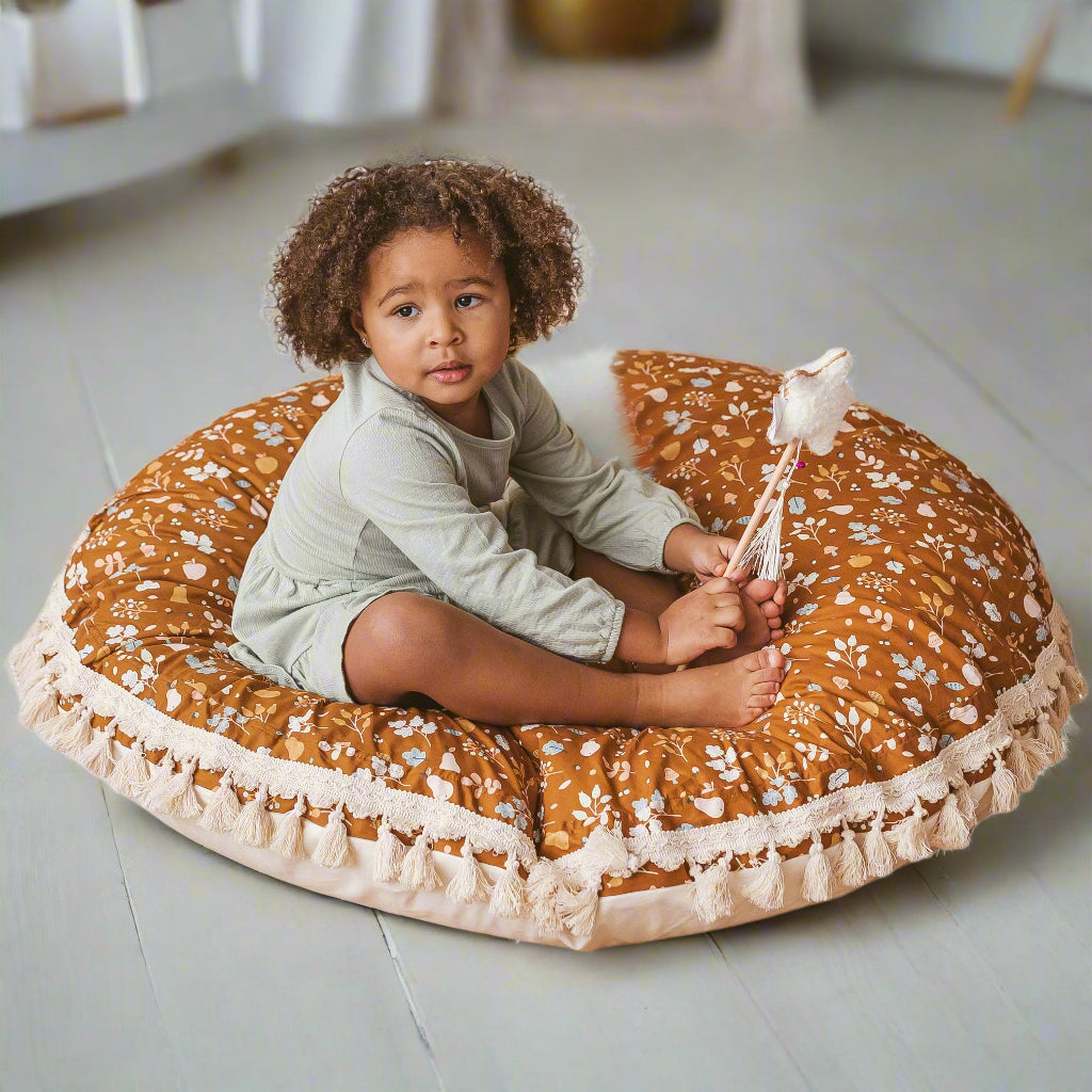 Child sitting on a round floral-patterned floor cushion which is brown with white flowers and a white trim with tassels
