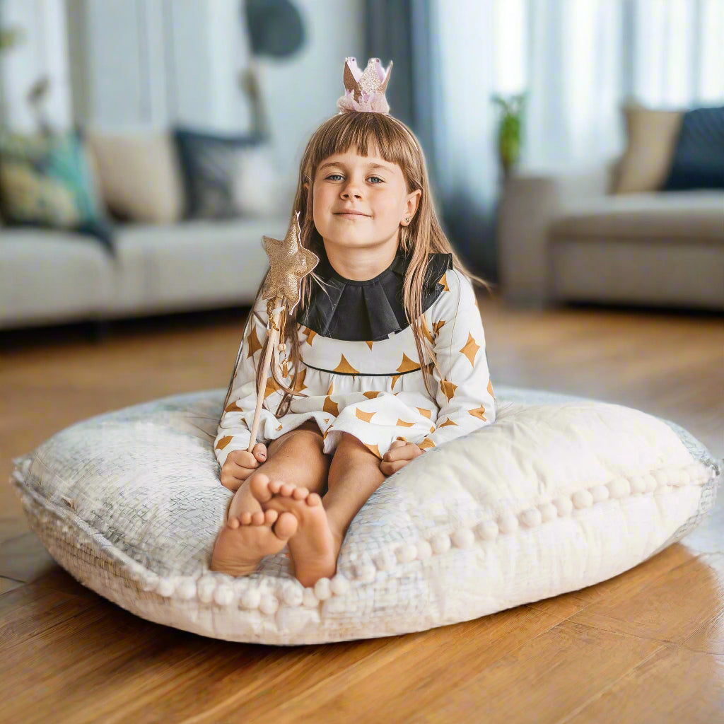 Child sitting on a large cream coloured floor cushion with a pom pom trim