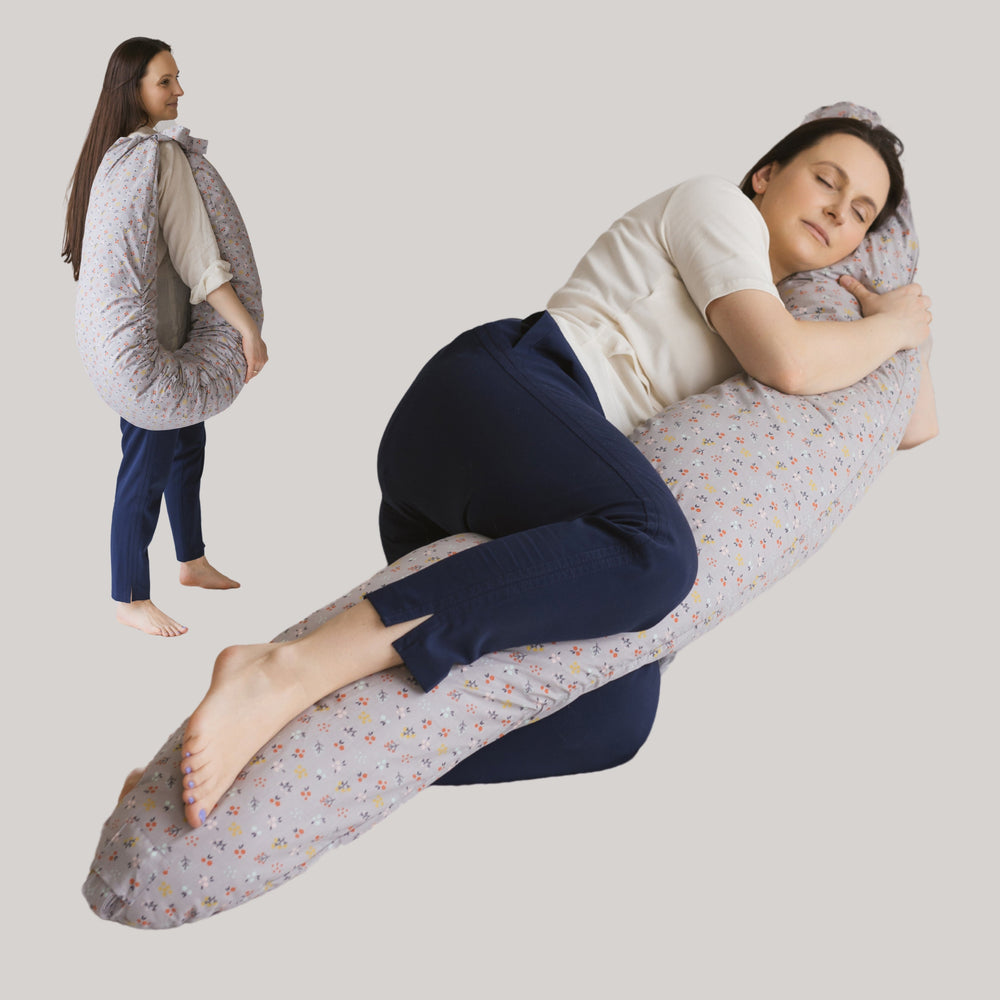 Woman lying down using a large support pillow which is fully extended to the length of her body and her leg wrapped over it. In background is another image of the same woman standing up with the pillow shared into its C shape and tied together with straps in a bow over her shoulder