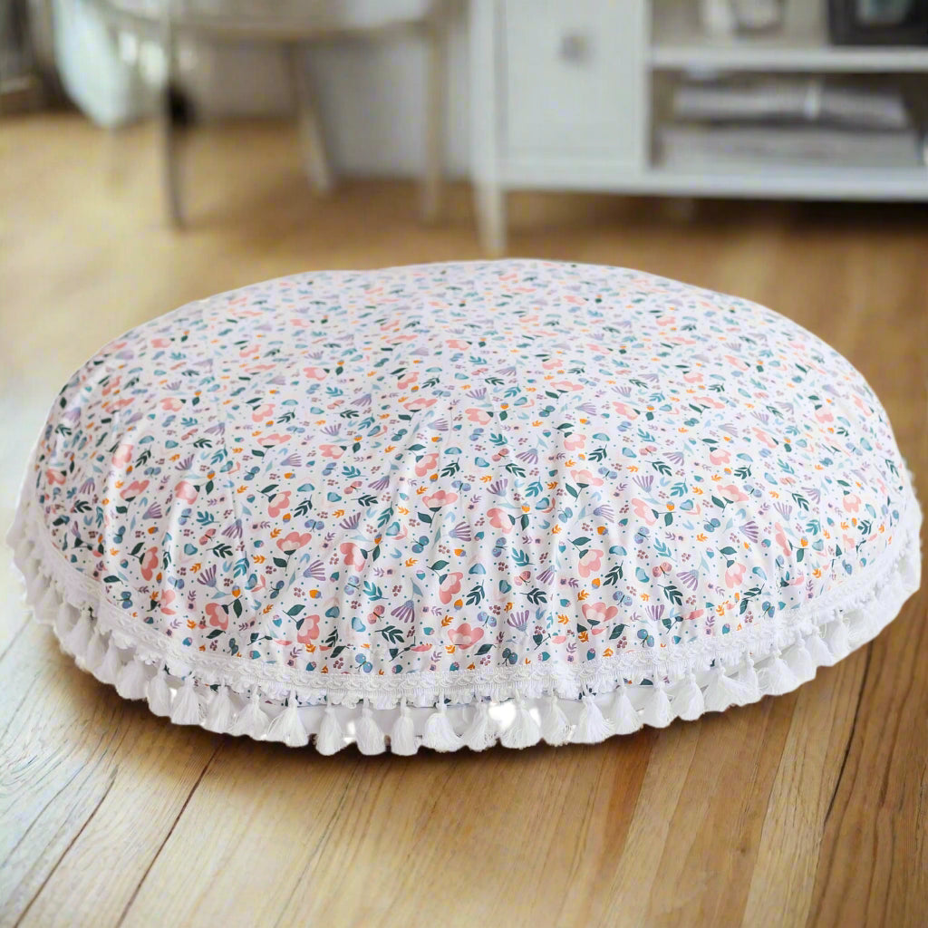 Large round floor cushion with a white background and a detailed floral design with tassels as a trim 