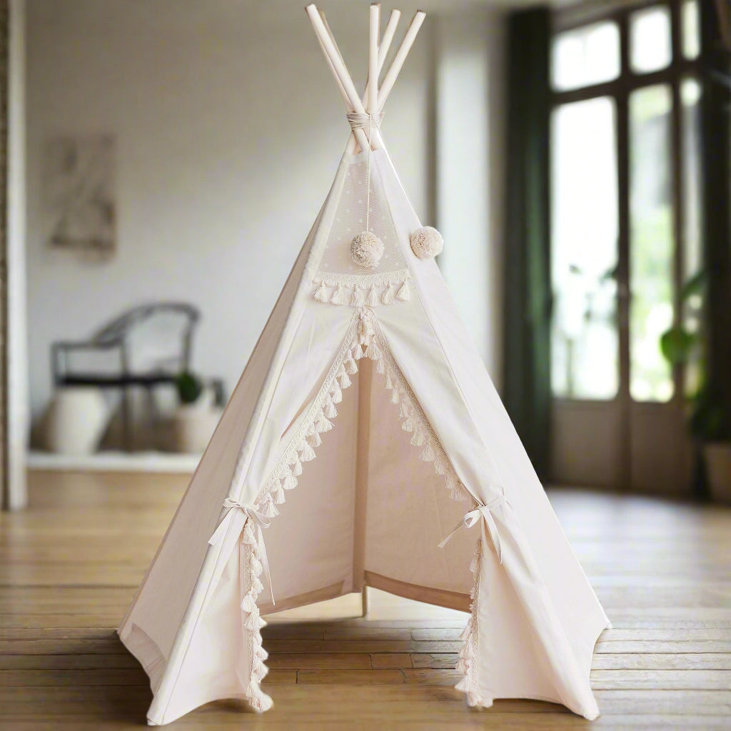 White teepee tent with white poles crossing over at the top, with round pom-poms hanging from the poles and white trim with tassles on the edge of the tent opening, which is tied open