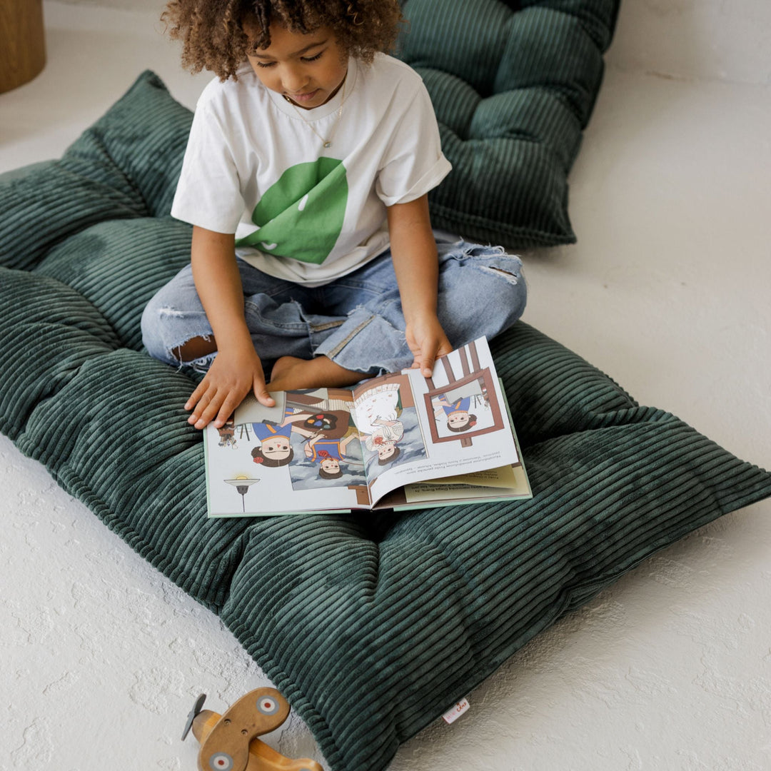 Child reading a book on a green cushioned floor pillow.