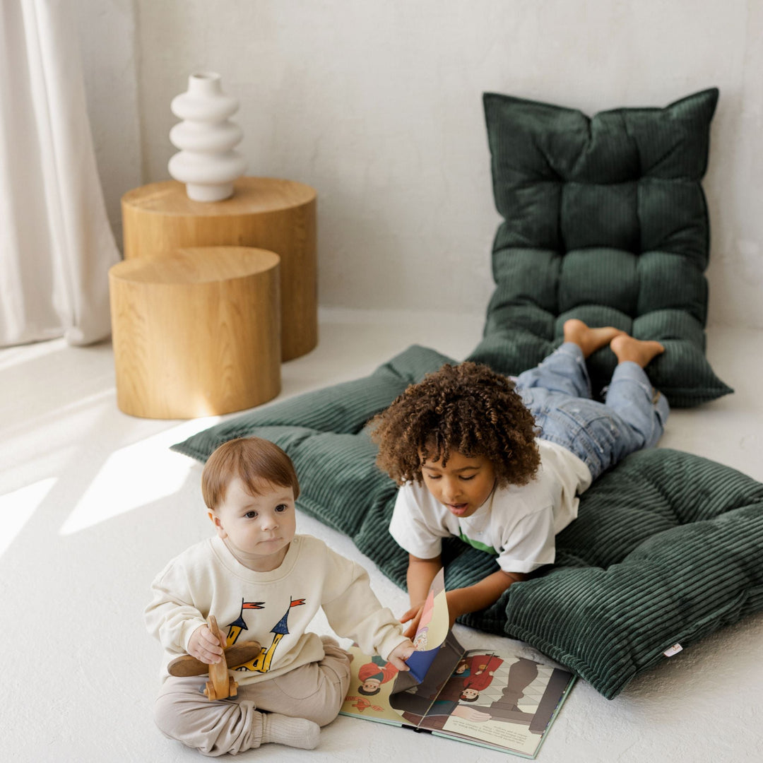 Two children sitting on green cushions reading a book in a room with wooden tables and a white wall.
