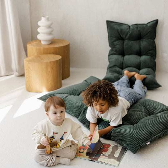 Two children sitting on green cushions reading a book in a room with wooden tables and a white wall.