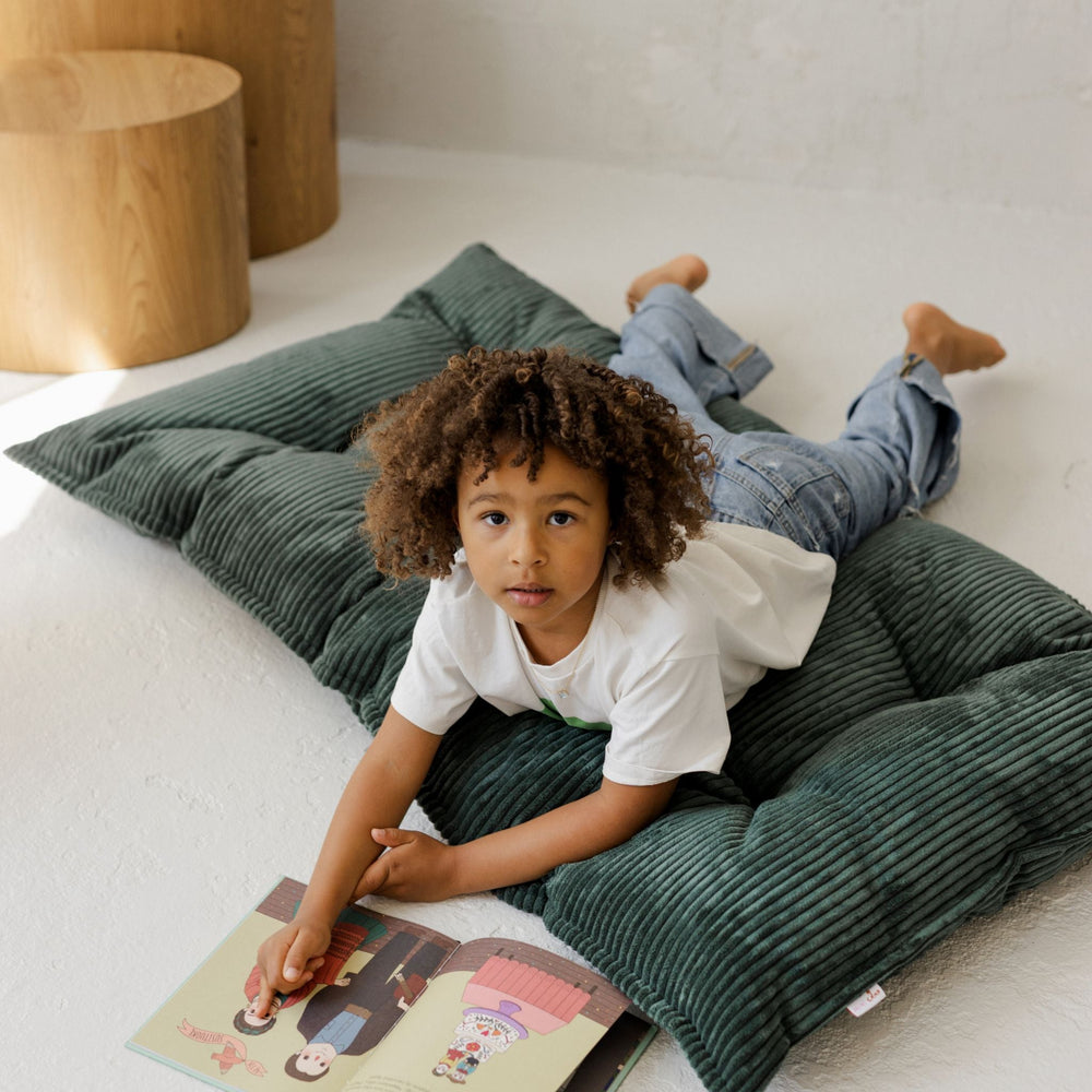 Child lying on a green cushion reading a book indoors