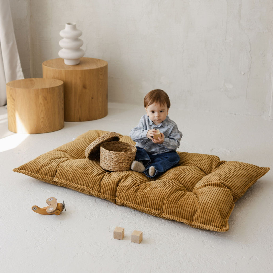 Young toddler sitting on a mustard yellow cushion and holding a toy in a minimalistic room with wooden furniture and toys.
