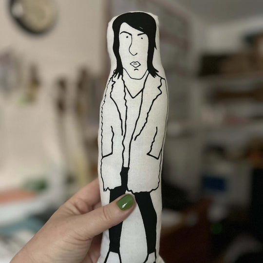 Hand holding screen printed doll cushion of Noel Fielding up against out of focus background