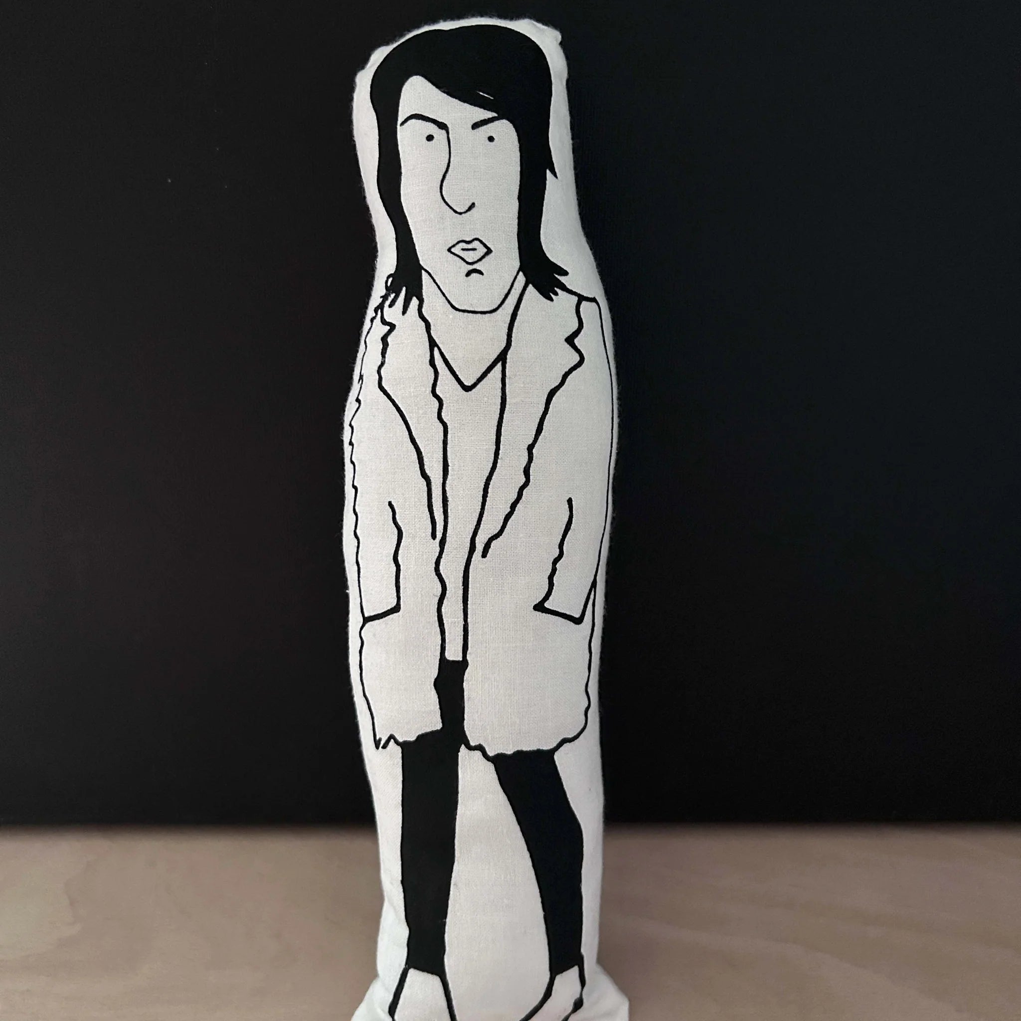 Screen printed doll cushion with white fabric and black illustration depicting TV presenter and comedian Noel Fielding, wearing black skinny jeans, heeled boots and an oversized coat