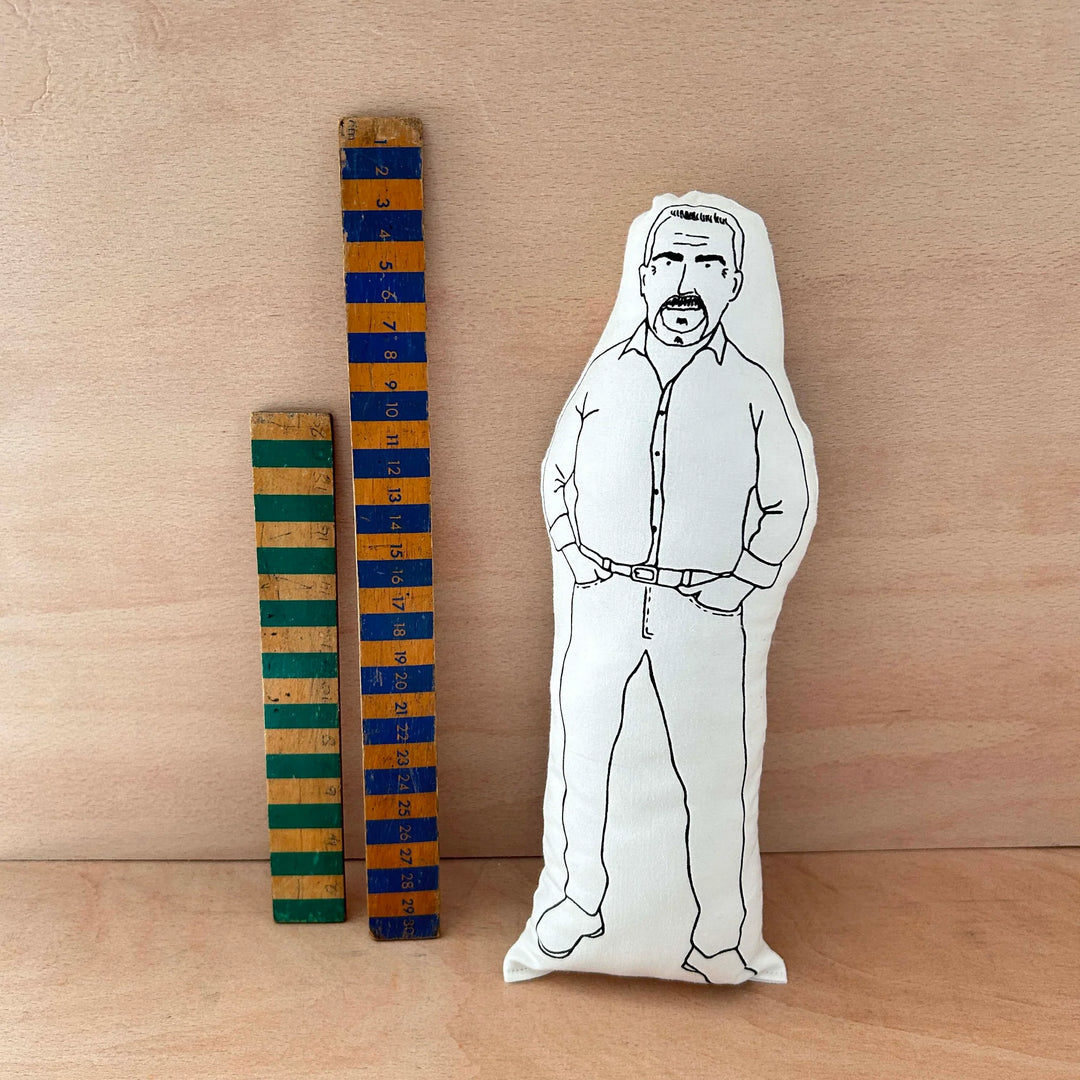 Screen printed doll cushion of Paul Hollywood next to two wooden rulers on a wooden background