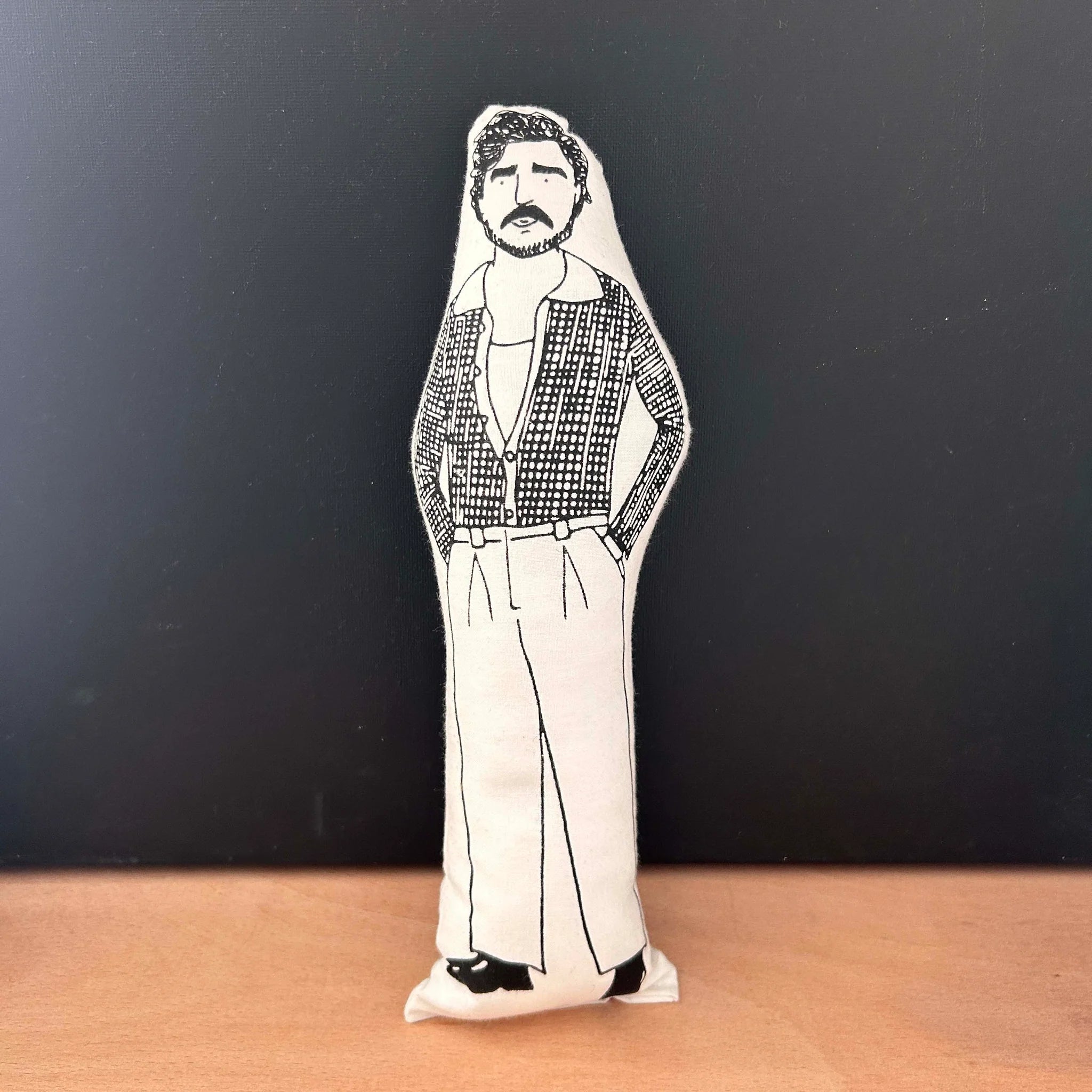 Screen printed doll cushion with white fabric and black illustration depicting Pedro Pascal wearing smart trousers, black shoes and an open checked shirt with his hands in his pockets
