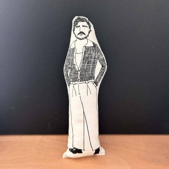 Screen printed doll cushion with white fabric and black illustration depicting Pedro Pascal wearing smart trousers, black shoes and an open checked shirt with his hands in his pockets