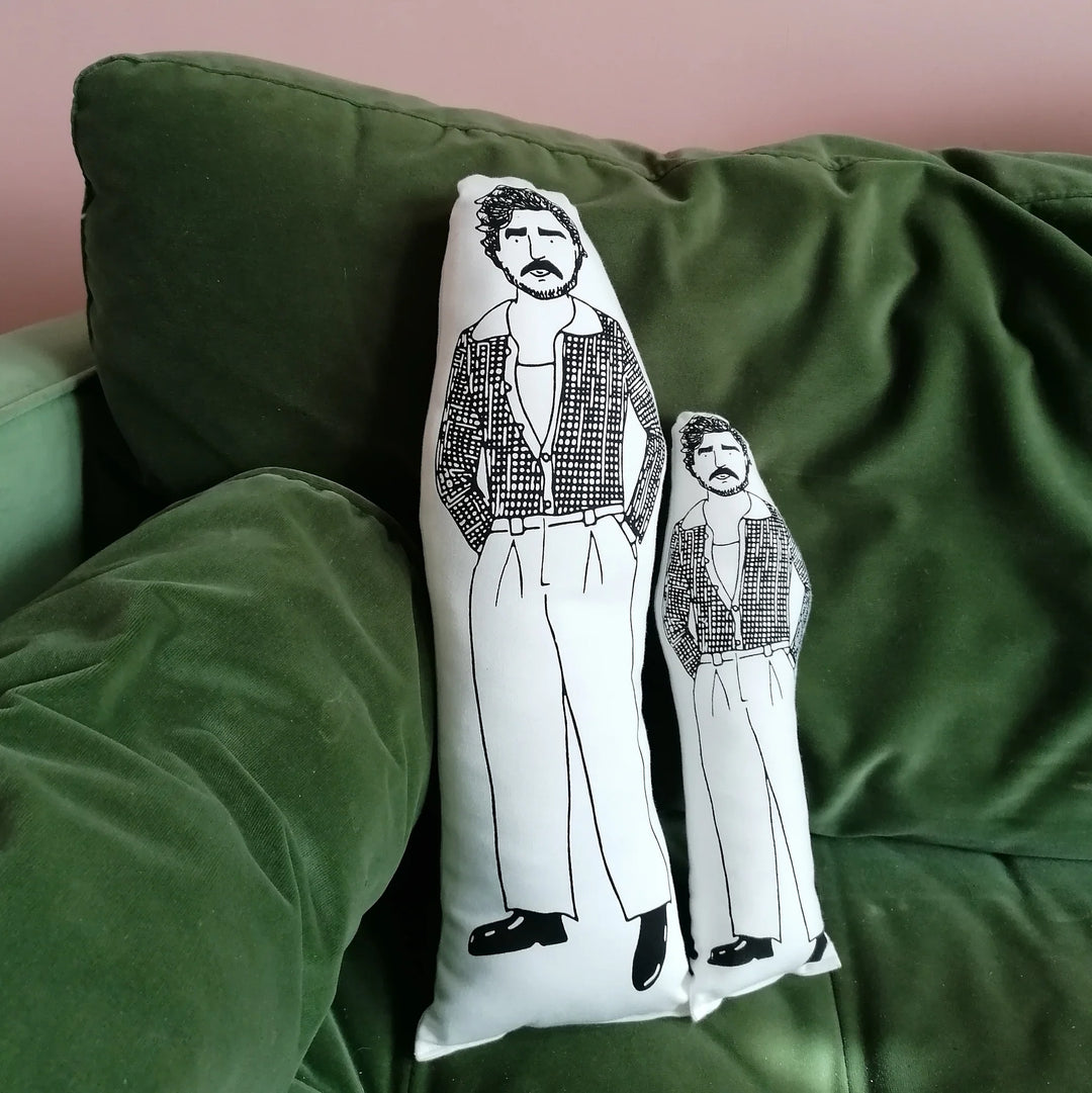 Two different sizes of the screen printed Pedro Pascal doll cushions next to each other on a green velvet sofa