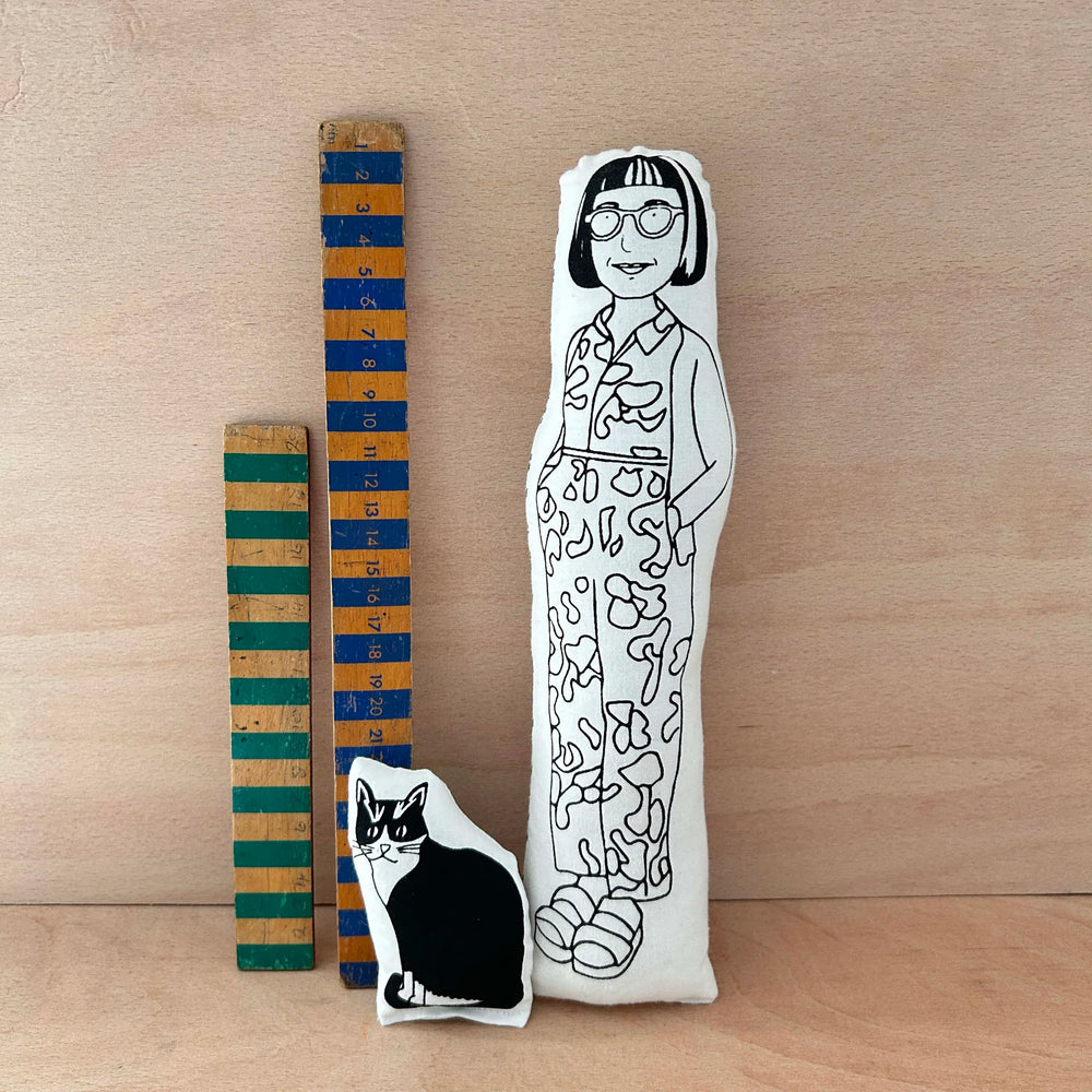 Two screen printed doll cushions of Philippa Perry and her cat Kevin next to two wooden rulers on a wooden background