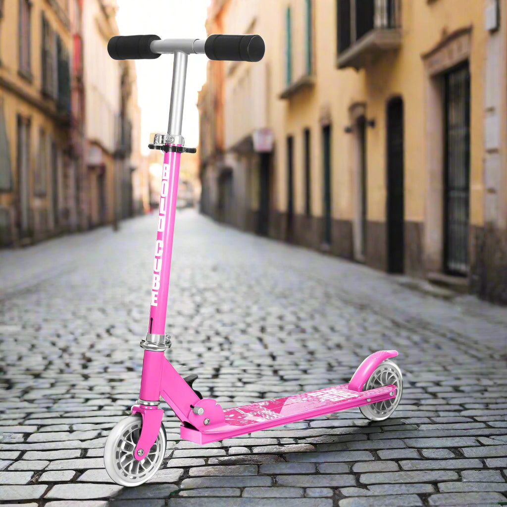 Pink scooter with black foam handles and 'Bold Cube' written on the vertical bar on a cobbled pavement background