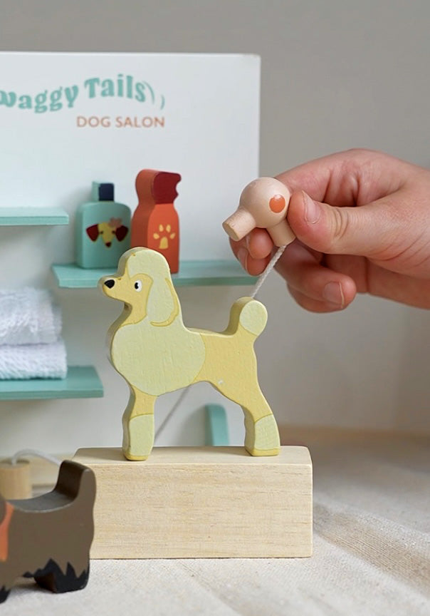 Wooden dog toy on a stand with a hand pulling a string, set against a background of 'Waggy Tails Dog Salon'.