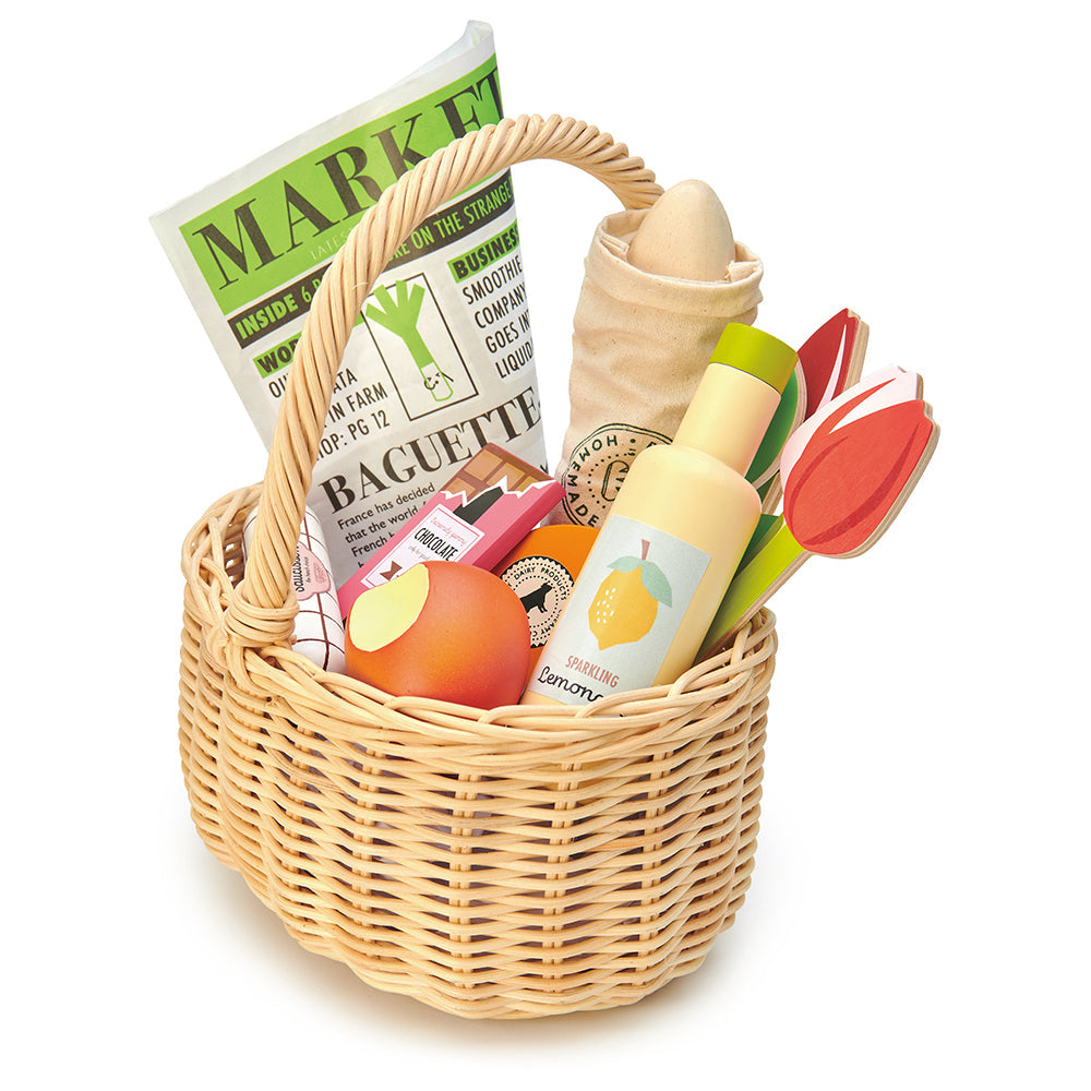Toy wicker basket with groceries including a newspaper, bread, flowers and fruits on a white background