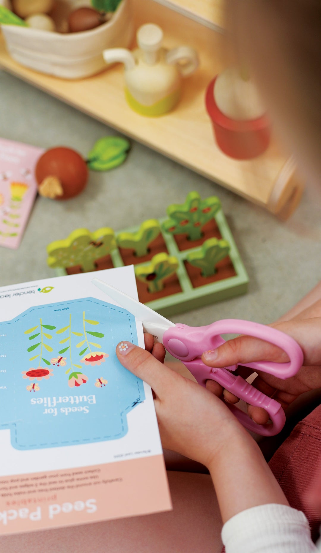 Child's hand holding pink scissors near a book with a garden illustration on a play mat with toys.