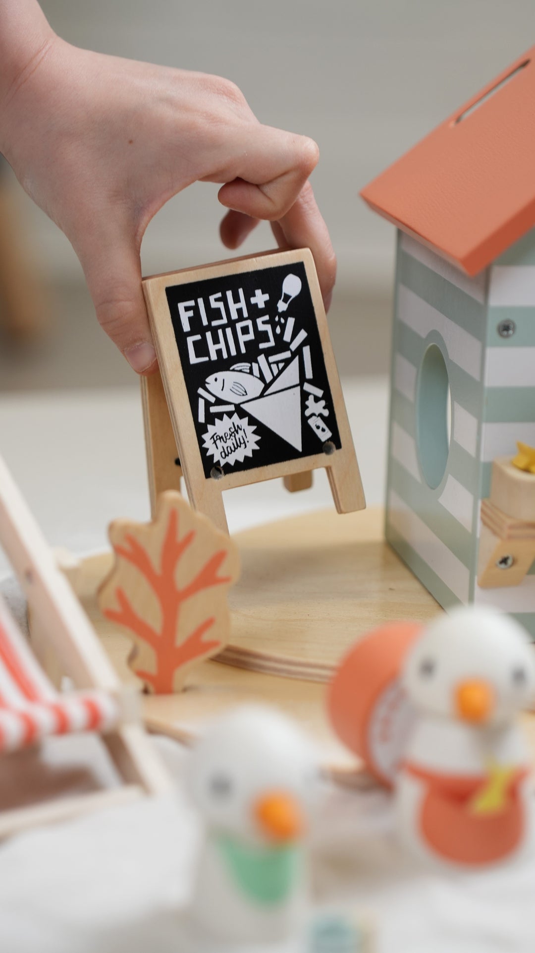 Hand holding a small wooden sign with 'Fish & Chips' text in a playroom setting.
