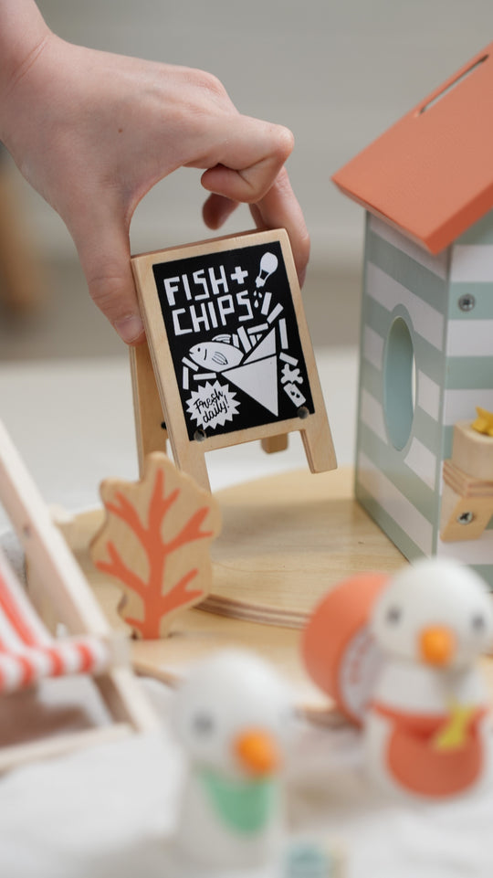 Hand holding a small wooden sign with 'Fish & Chips' text in a playroom setting.