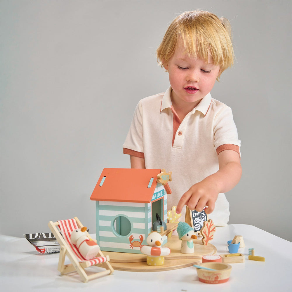 Child playing with a wooden toy hut and accessories on a white surface.