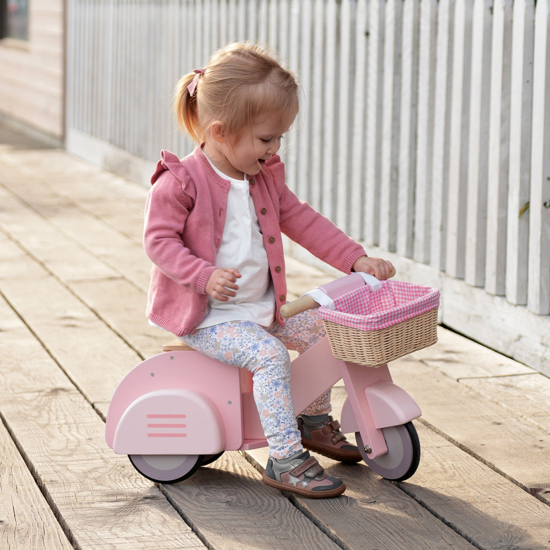 Child riding a pink toy scooter with a basket on a wooden deck.