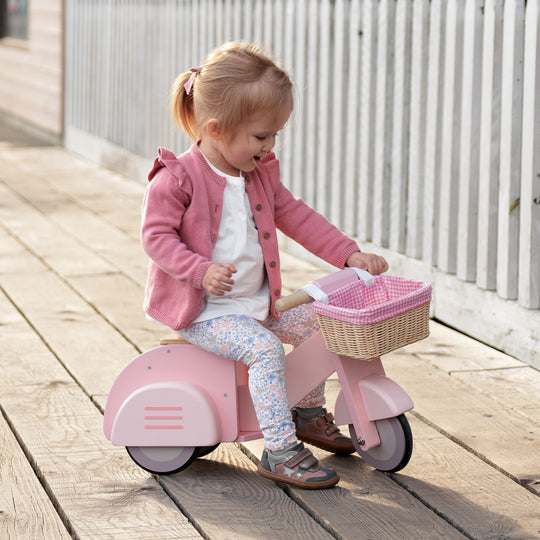 Child riding a pink toy scooter with a basket on a wooden deck.