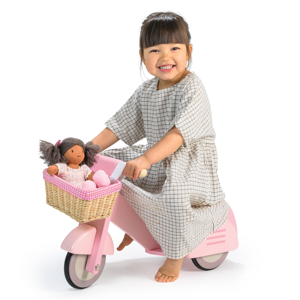 Child on a pink scooter with a basket containing a doll and teddy bear, wearing a checkered dress.