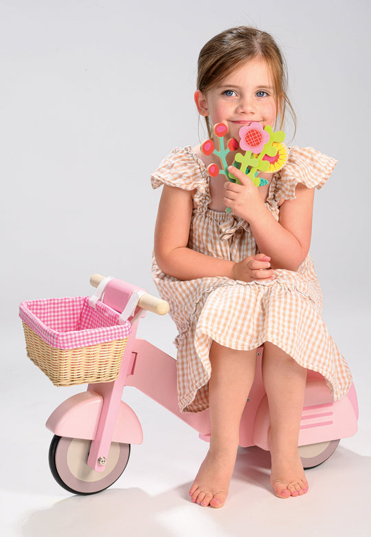 Young girl sitting on a pink toy motorcycle holding a colorful flower toy against a white background