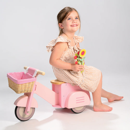 Young girl sitting on a pink tricycle holding flowers against a light grey background
