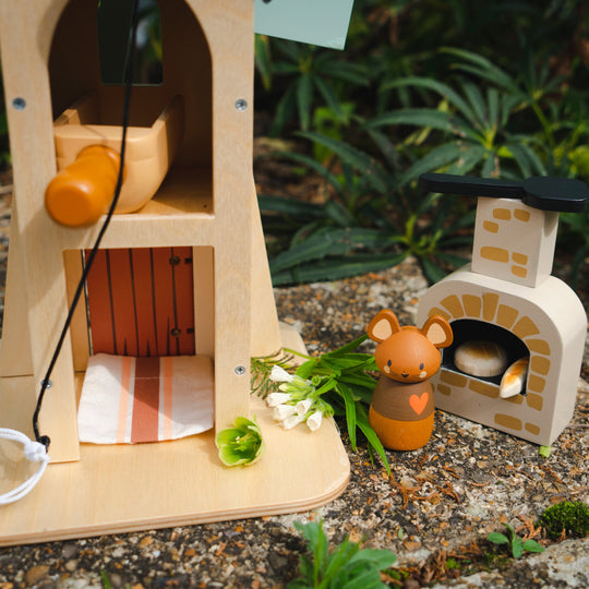 Small wooden mouse figurine next to a miniature house with plants and flowers outdoors.