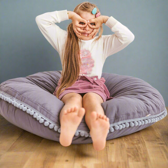 Child sitting on a round grey cushion with pom-poms