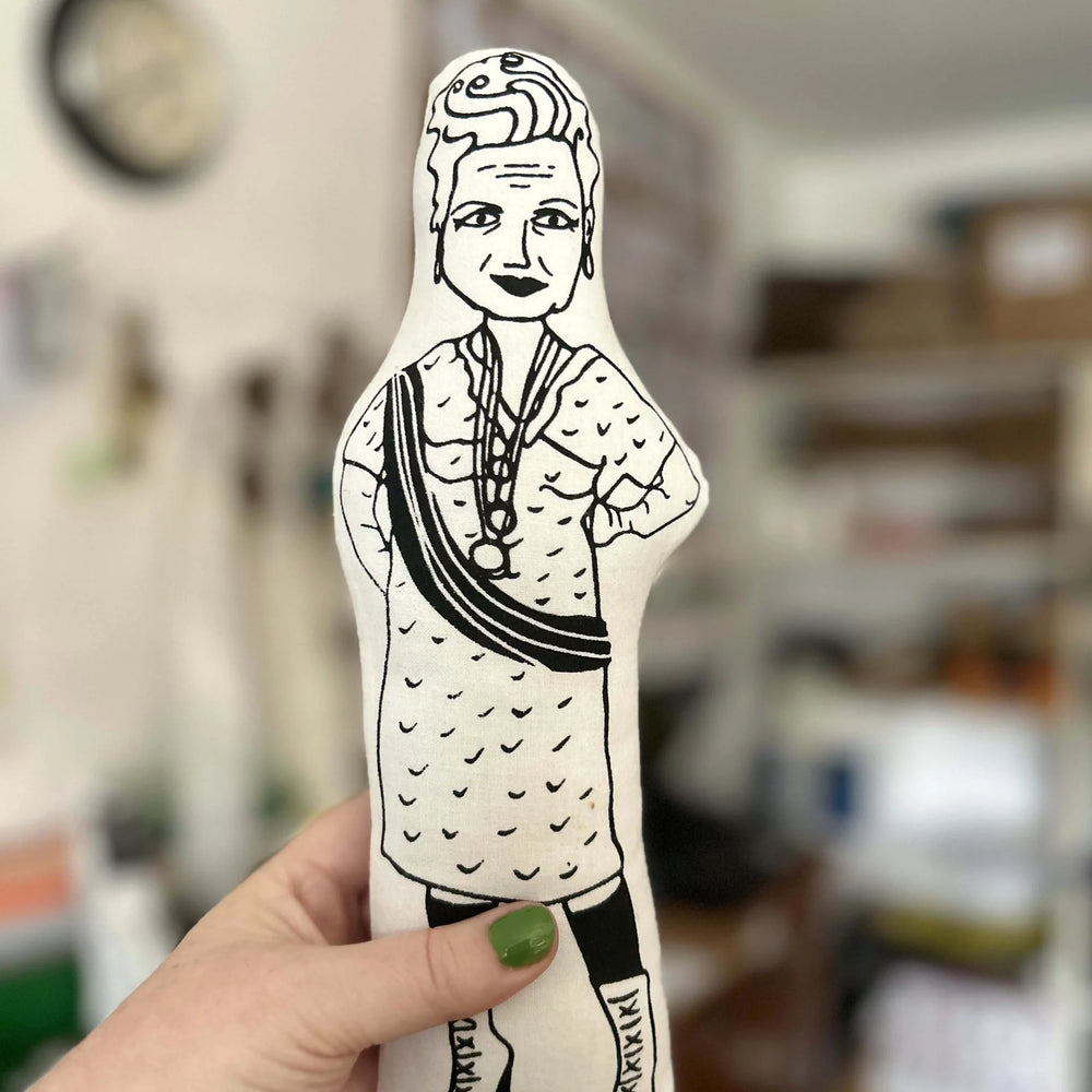 Hand holding a black and white illustrated figure of Vivienne Westwood against a blurred indoor background