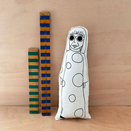 Decorative pillow with a black and white design of a person wearing sunglasses and polka-dotted dress, next to wooden rulers on a wooden surface.