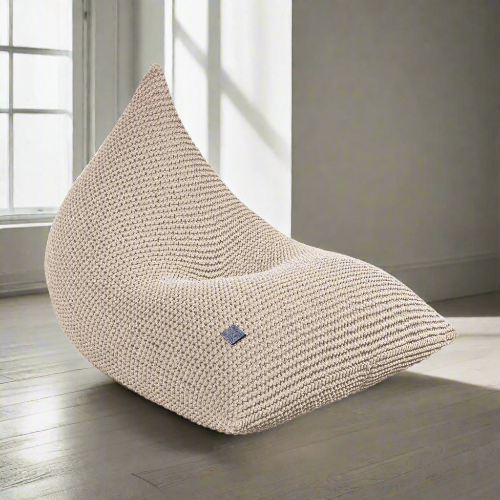A beige handmade knitted bean bag chair with a zipper, made of chunky cotton cord, with a small blue tag on the lower side.
