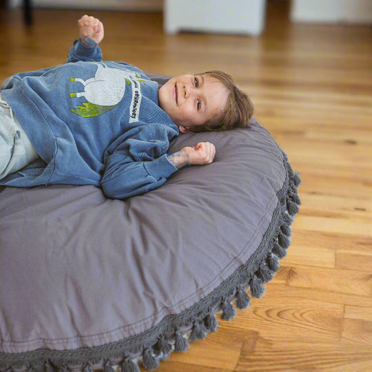 Child lying on a round grey cushion with tassels