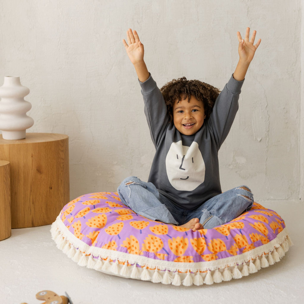 Child sitting on a colourful round cushion with raised arms, wearing a grey sweatshirt
